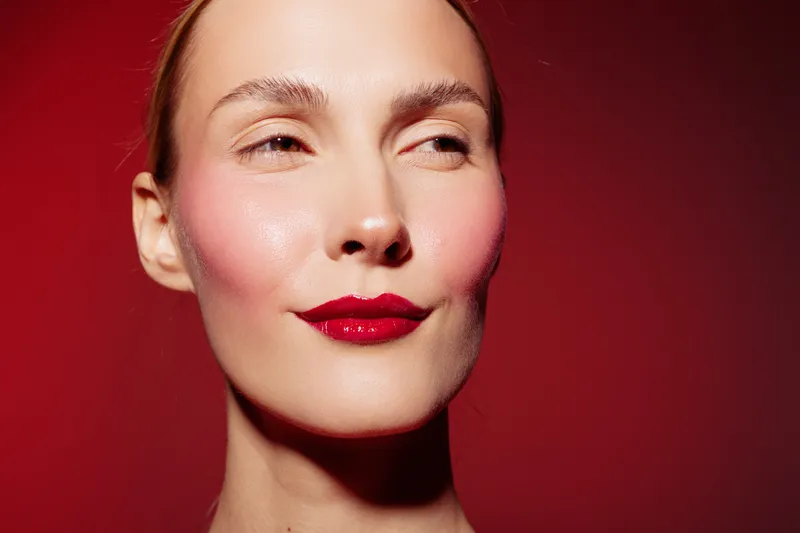 Close-up portrait of confident woman with red lipstick and glowing skin on red background. Concept of beauty, makeup, skincare, and self-confidence in studio lighting.