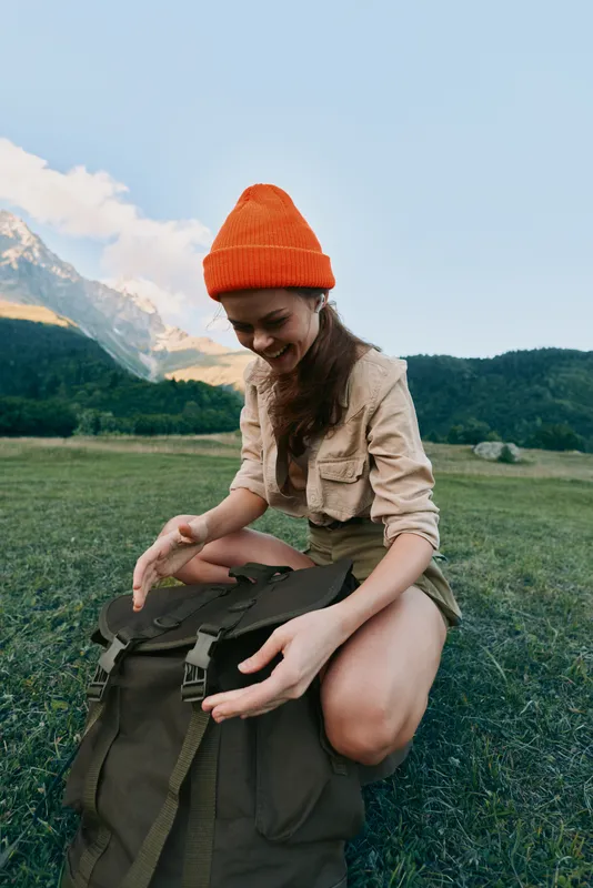 Woman in an orange beanie sits on grass beside a backpack in a mountain meadow, enjoying outdoor life and natural scenery and sunlight warms her face under a clear blue sky