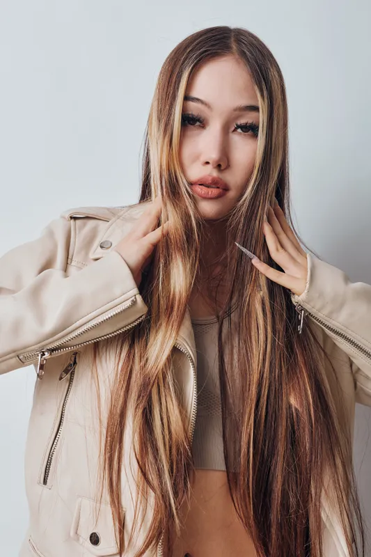 Young woman with long straight hair wearing beige jacket and crop top posing against white background for fashion portrait and beauty closeup indoors.