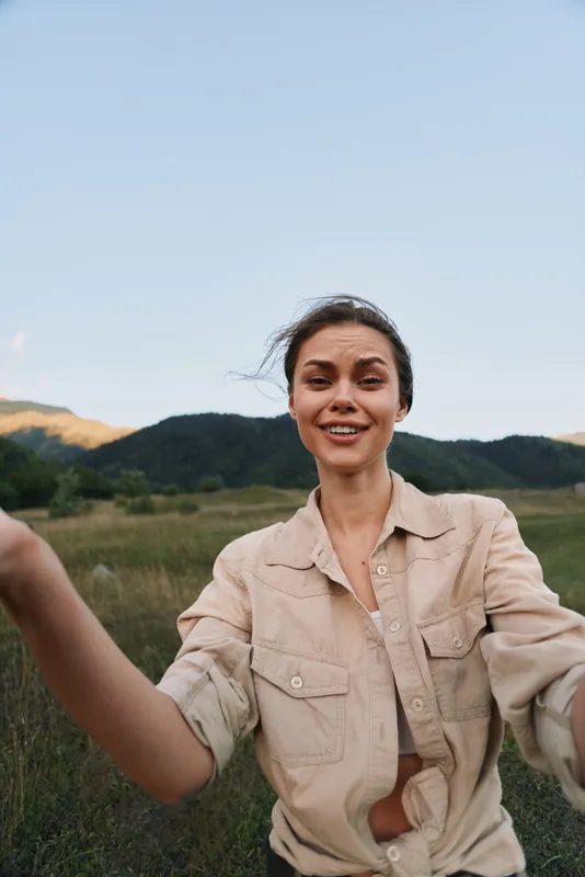 Young woman with dark hair wearing beige jacket smiling outdoors in a green field with hills and blue sky background, capturing lifestyle and nature concept.