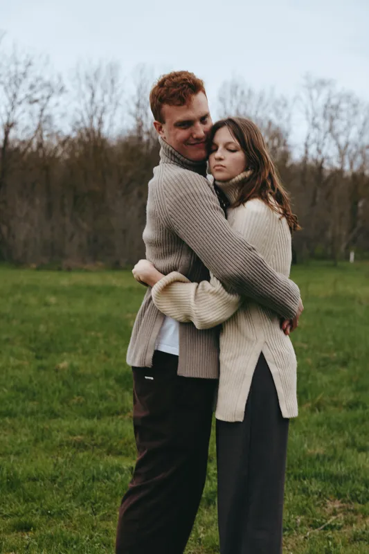 Two adults embrace in a cozy outdoor scene, a man and woman in knit sweaters share a warm moment in a grassy field, soft natural light highlights affection and romance, creating a candid lifestyle