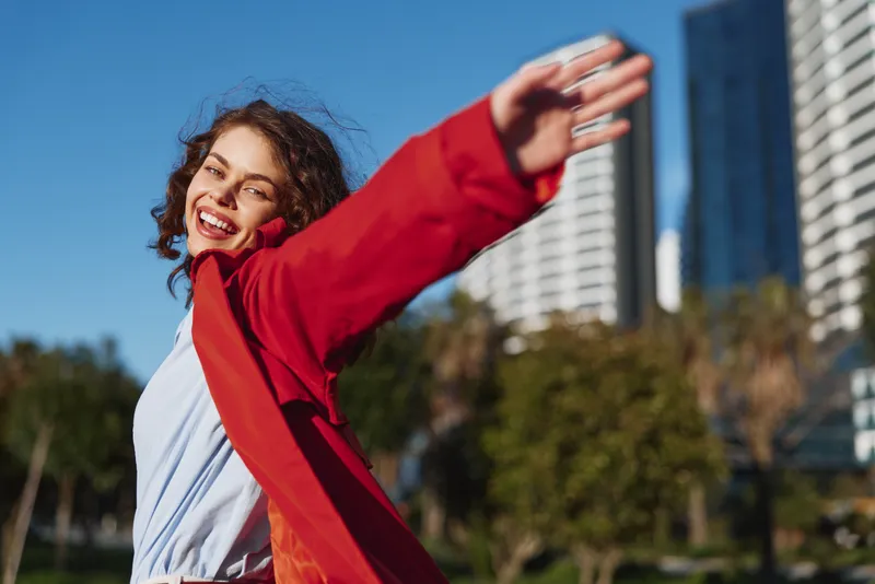 Cheerful woman outdoors wearing red jacket smiling in sunlit urban park with open arms and lively energy bright colors and natural light in a windy moment
