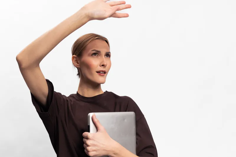 Young woman holding laptop raises hand to shield eyes from bright light. Casual clothing and neutral background create a simple, modern concept of technology and concentration.