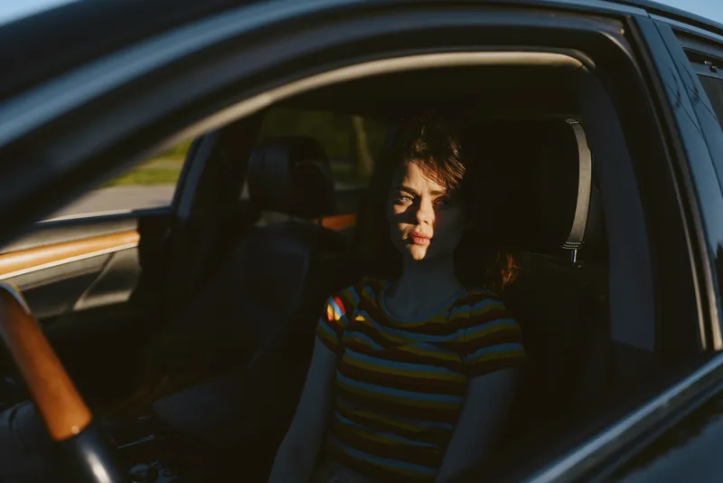 Confident woman inside a car, sunlit face, striped top, relaxed posture, warm retro film color, nostalgic mood, candid lifestyle moment, natural textures and muted light hints, soft.