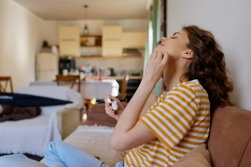 Smiling woman in striped shirt applying makeup at home, relaxed lifestyle moment, soft natural light, warm living room ambiance, focus on beauty, skincare, routine and selfcare.