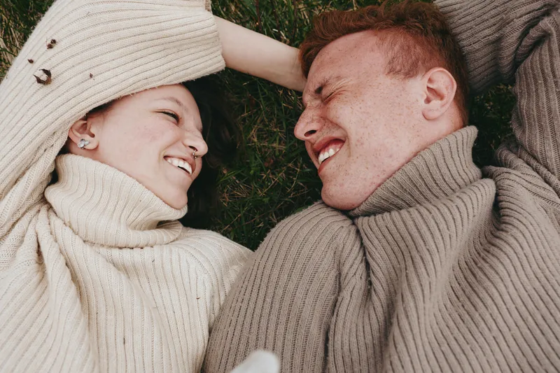 A couple lying on grass wearing sweaters, smiling and enjoying a cozy outdoor moment. A lifestyle scene of warmth, affection, and togetherness in a sunlit field.