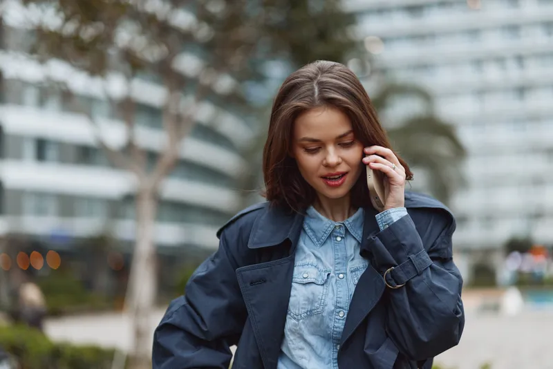 A woman in a denim shirt and dark jacket talks on her phone in an urban outdoor setting, showcasing casual style and everyday communication in a modern city.