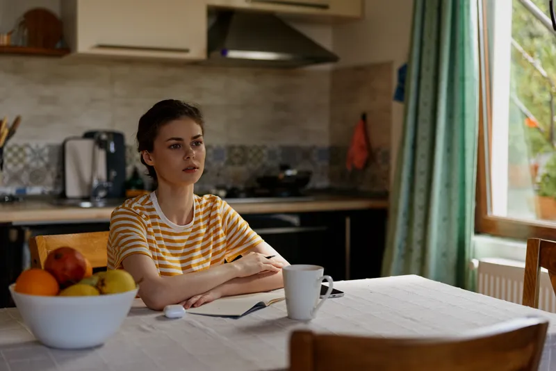 Calm woman sits at a kitchen table with a notebook, fruit, and mug, focused on a task. Natural daylight from a window creates a warm, cozy home mood and everyday lifestyle.