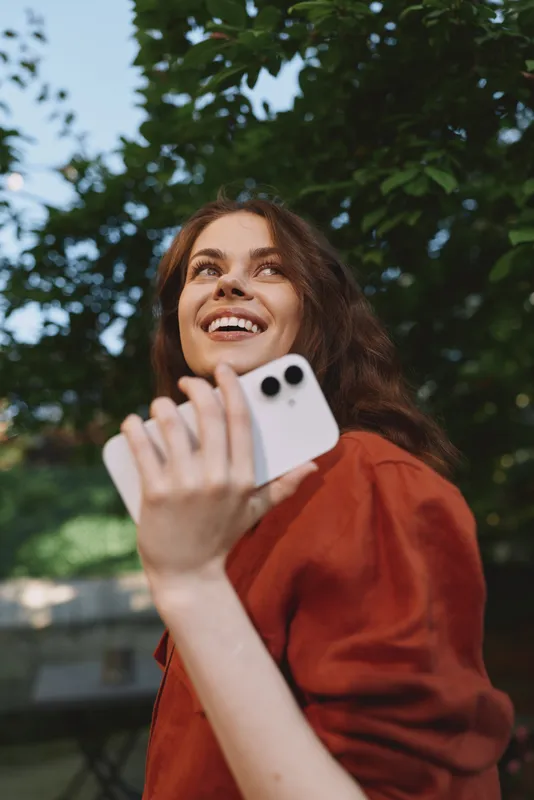 Candid woman lifestyle scene outdoors, smiling while holding a smartphone, casual red top, sunny park background, joyful moment captured in natural light and warmth