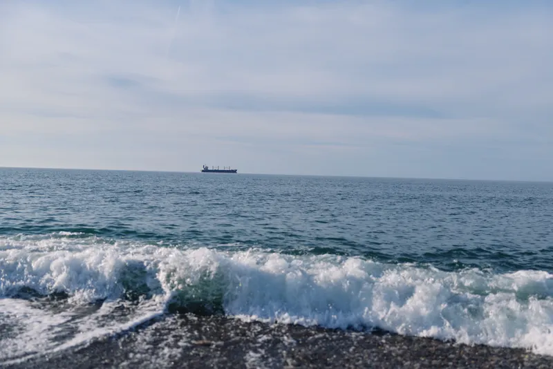 Ocean and sea meet at a tranquil shoreline with rolling waves, a distant ship on the horizon, a calm sky reflecting on the water and a pebbled shore in the foreground.