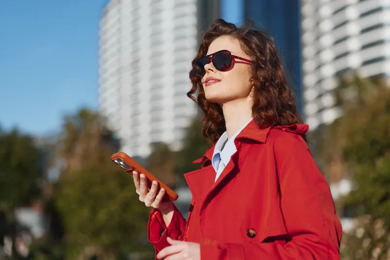 A stylish woman in a red coat wears sunglasses, looking up, holding a smartphone in a sunny urban setting with modern buildings behind, conveying confidence.