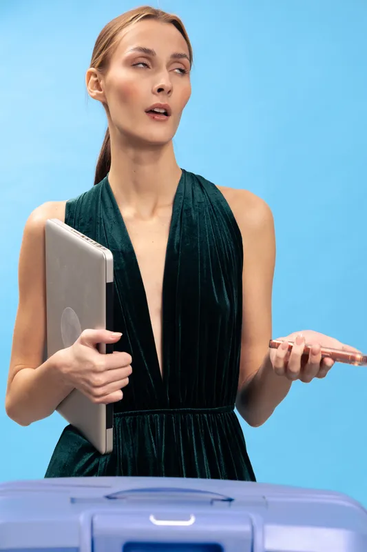 Young woman in dark dress holds laptop and phone, standing behind suitcase on blue background. Concept for travel, technology, business, and planning with thoughtful expression.