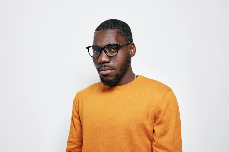 African American man with dark skin wearing glasses and orange sweater standing against white background. Portrait of confident young black male with short hair and beard in casual clothing.
