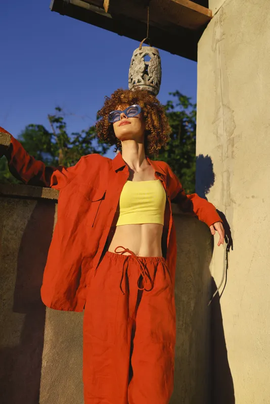 Confident woman wearing a bright red suit and yellow crop top stands on a sunlit terrace, posing with an outstretched arm. Fashion lifestyle mood with bold colors and chic streetwear