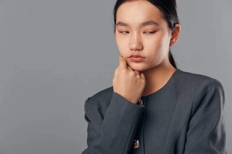 Young Asian woman with long dark hair wearing gray jacket posing with thoughtful expression and hand on chin isolated on solid gray background in studio portrait.