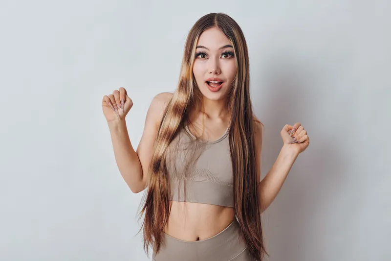 Young woman with long hair in casual activewear posing surprised and excited with open mouth and raised hands on light neutral background indoors.