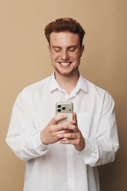Smiling young man with curly hair wearing white shirt looks at smartphone with closed eyes. Studio portrait on beige background. Concept of technology, communication, and happiness.