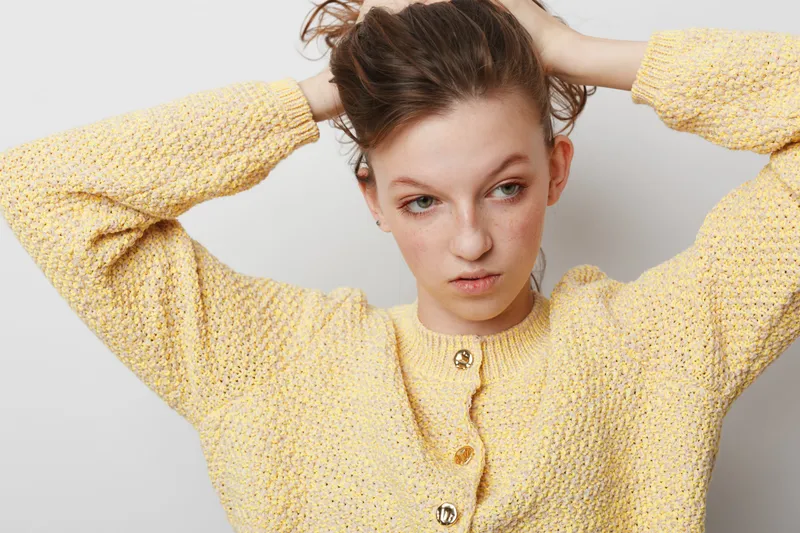 Young woman in yellow knitted sweater adjusts her hair with both hands, looking down thoughtfully. Studio portrait on white background with soft natural lighting and casual style.