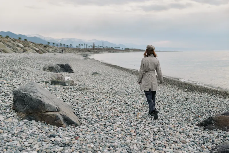 Woman in beige coat and hat walks alone on rocky beach near calm sea under cloudy sky. Concept for solitude, nature, travel, and peaceful outdoor lifestyle in cool weather.