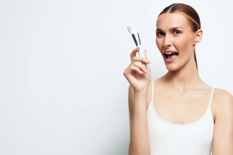 Young woman in white tank top holds two makeup brushes near face smiles with open mouth on white background beauty concept skincare routine