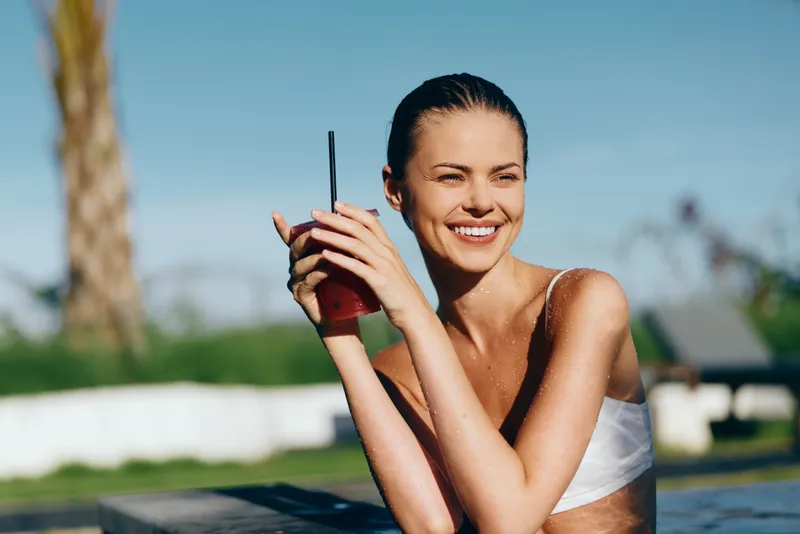 Smiling woman with wet hair in a white bikini holds a cold beverage by a poolside, bright outdoor scene, wellness and lifestyle vibe, sunlit mood, relaxed and refreshing.