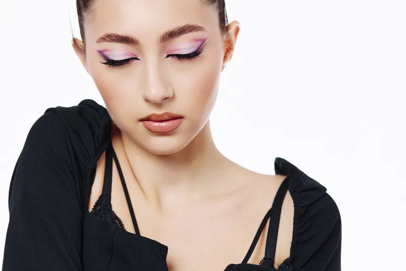 Young woman with creative pink and purple eyeshadow posing in a black outfit on white background. Concept of beauty, fashion, and avant-garde makeup fashion photography.