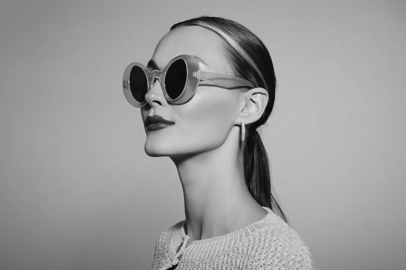 Profile of a fair skinned white woman wearing round sunglasses and earrings, styled hair pulled back, dressed in a textured top, isolated on white background.