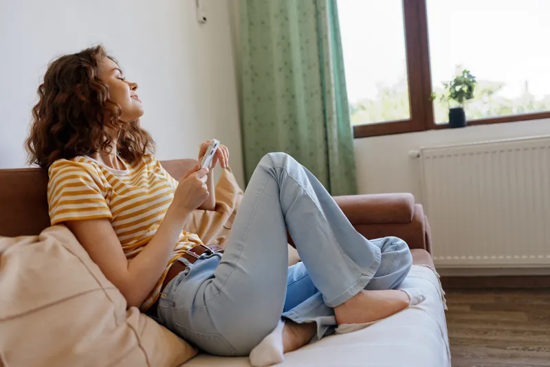 Casual woman relaxing at home sits on a sofa wearing a striped tee and jeans, enjoying downtime with a handheld device, bright natural light, calm domestic mood.