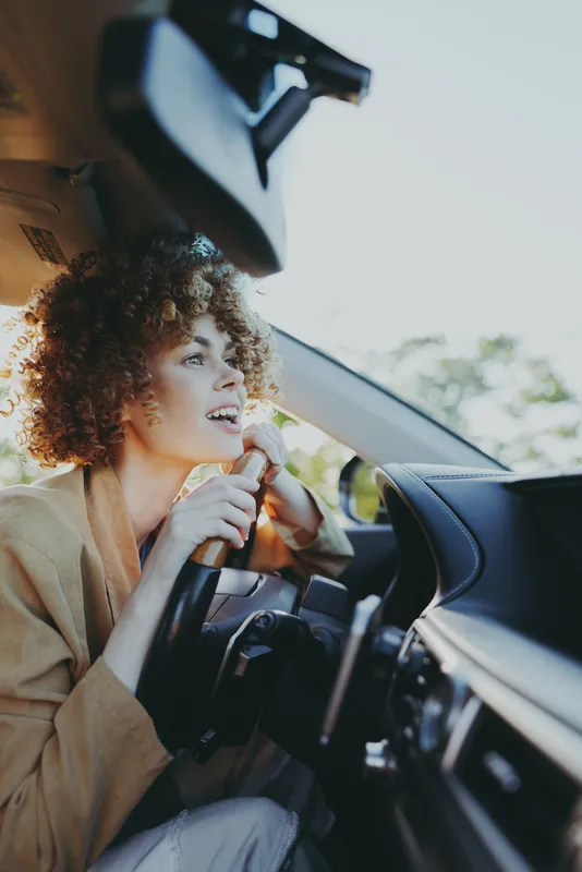 A woman driver sits behind the wheel, smiling with joy in the bright car interior. A candid travel moment highlighting lifestyle, optimism, and everyday driving.