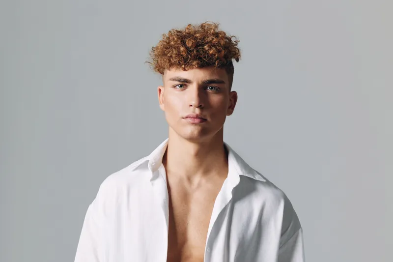 Young man with curly hair wearing open white shirt posing against gray background, looking confidently at camera with neutral expression and relaxed posture.