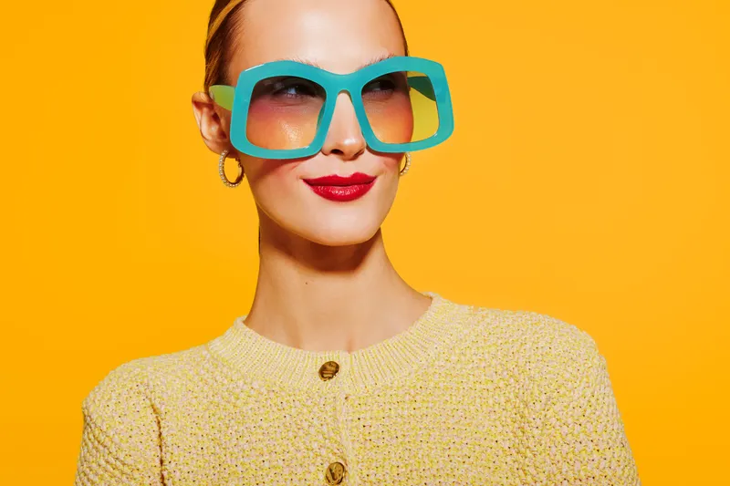 Stylish woman with large turquoise sunglasses and red lipstick poses against bright yellow background. Fashion concept with vibrant colors and confident expression in studio lighting.