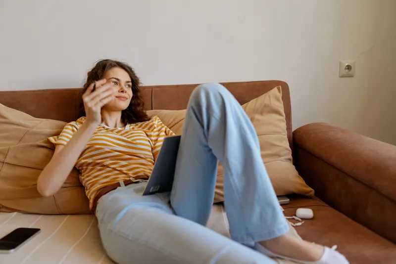 Casual woman lounging on a sofa at home, using a laptop with a relaxed posture. Warm natural light creates a cozy livingroom mood for everyday work and leisure.