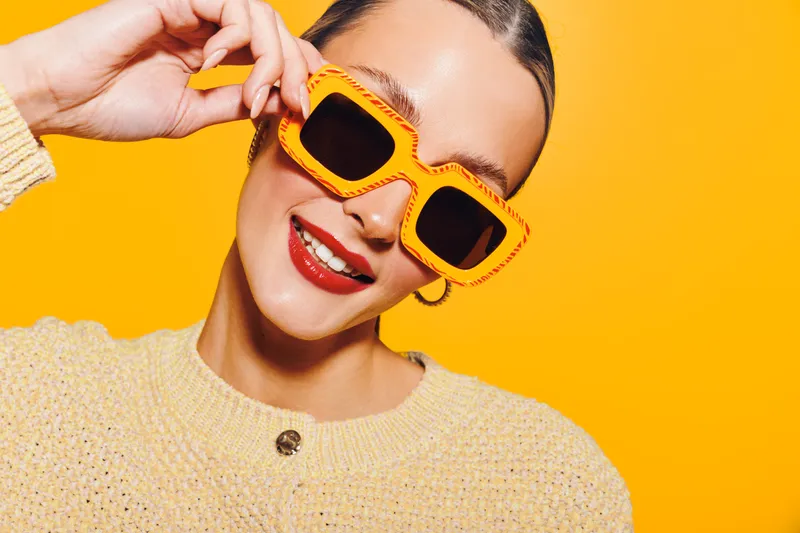 Young woman with bright orange sunglasses and red lipstick smiles confidently on yellow background. Fashion concept with vibrant colors and stylish accessories for summer look.