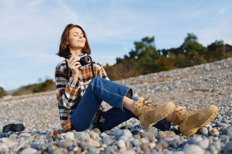 Woman photographer on a rocky beach smiles with a camera, relaxed pose under blue sky, capturing outdoor scenery and nature during a casual photo session.