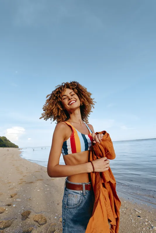 Smiling woman with curly hair wearing striped bikini top and jeans holding orange jacket on sandy beach near calm sea under clear blue sky. Summer vacation concept.
