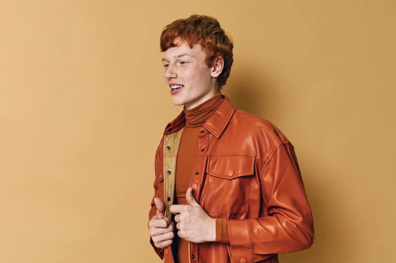 Young man with red hair wearing a brown leather jacket and turtleneck posing against beige background in studio for fashion or portrait concept with casual style and confident expression.