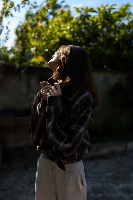 Brunette woman in a plaid shirt stands outdoors, tilting her head up toward the sun as she gazes skyward with relaxed hands near her chest
