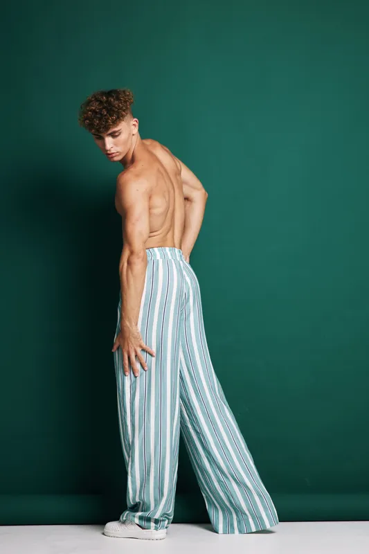 Young man with curly hair posing shirtless in striped wide leg pants and white sneakers against green background. Fashion, style and casual outfit with confident posture.
