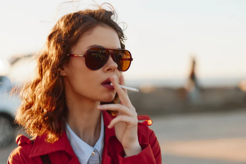 Cheerful woman outdoors, sunglasses on, red jacket, smoking cigarette, sunlit moment, relaxed posture, urban waterfront background, warm tones