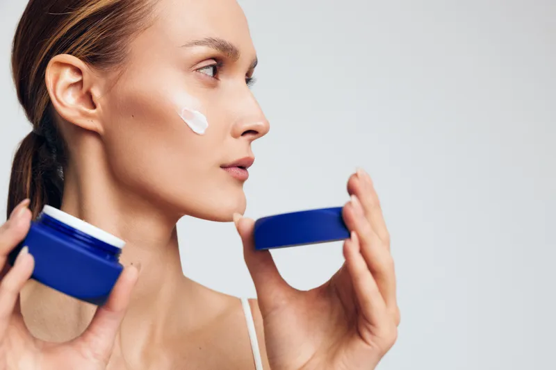 Young woman applies moisturizing cream on face holding blue jar with white lotion isolated on gray background skincare beauty routine daily care concept.