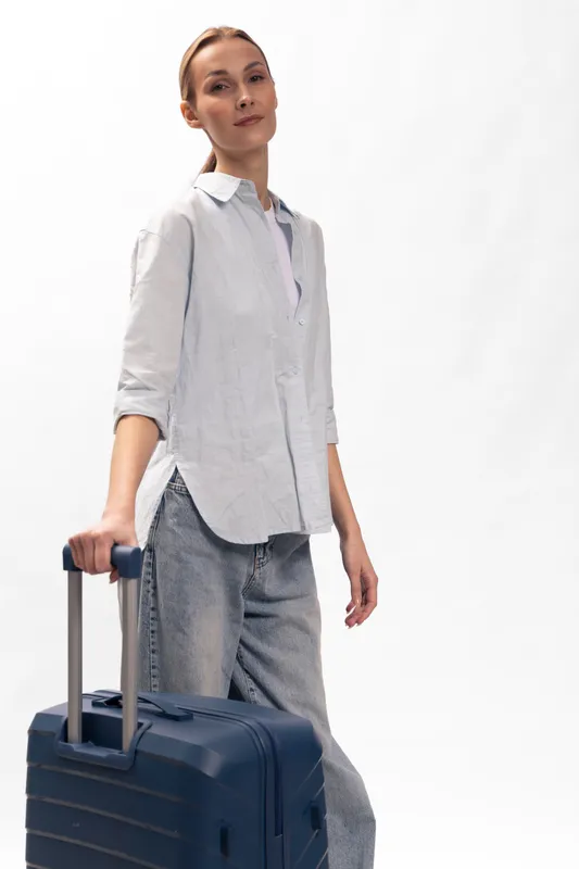 Young woman in casual shirt and jeans stands with dark blue suitcase on white background. Concept for travel, tourism, lifestyle, and modern journey preparation.