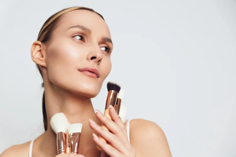 Young woman with clear skin holding makeup brushes close to face, looking sideways, minimal makeup, soft lighting, simple white background, beauty concept