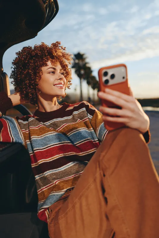 Woman lifestyle portrait outdoors, curly hair, striped sweater and brown jacket, holding smartphone selfie while sitting by car, golden sunset light, natural smile, casual travel moment