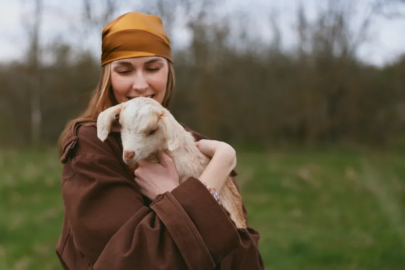 Young woman in a brown coat and orange headscarf gently embraces a small white baby goat outdoors in a natural green field during a calm overcast day.