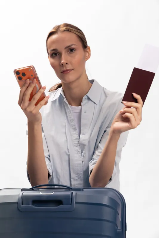 Young woman holding passport and smartphone stands behind suitcase, looking focused. Concept of travel planning, business trip, and modern communication with digital devices.