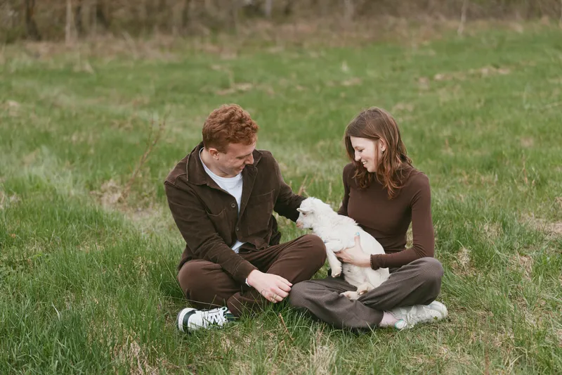 Young couple sitting on grass, playing with a small white dog in an outdoor setting. Casual lifestyle scene focused on companionship, nature, and bonding. Soft natural light creates a warm, intimate