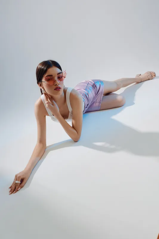 Young woman in stylish sunglasses and pastel outfit poses lying on white floor. Concept of beauty, fashion, and modern lifestyle with soft natural lighting.