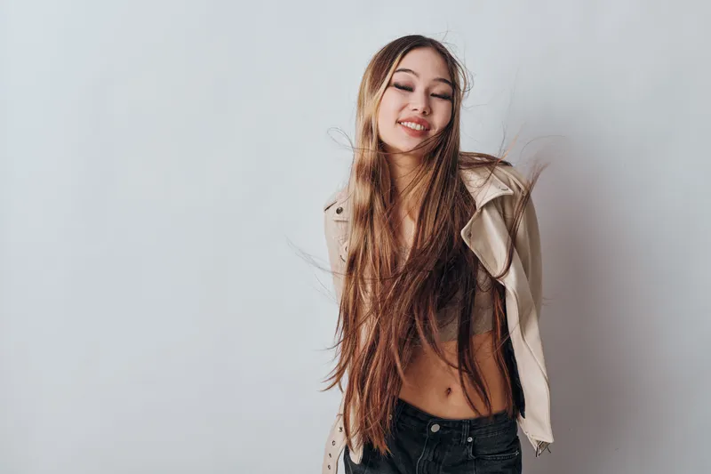 Young woman with long hair smiling with closed eyes against white background wearing open light jacket and black pants casual style and natural pose.