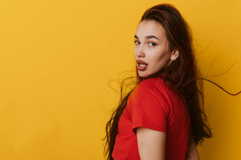Young woman with long hair wearing red t shirt posing against yellow background looking over shoulder with confident expression and natural makeup in studio portrait.