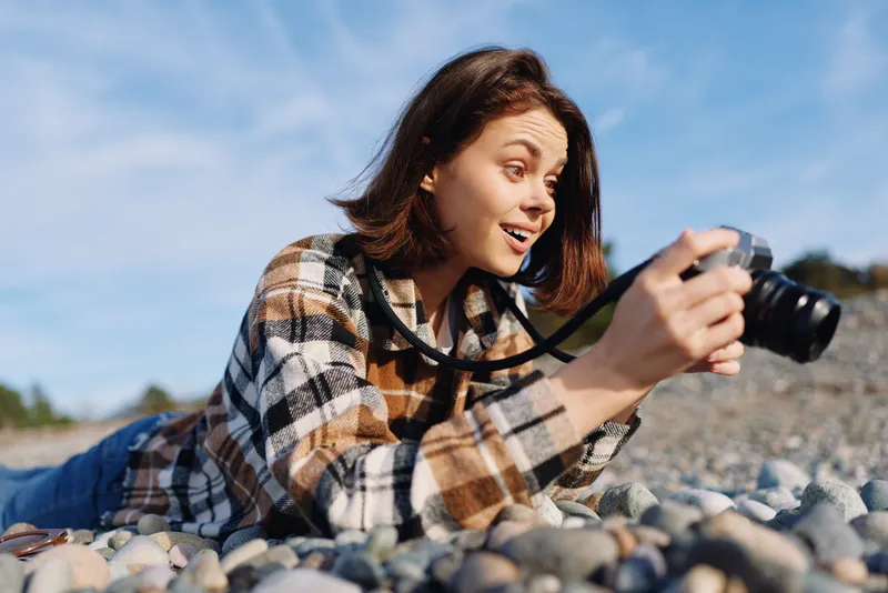 Woman photographer on a rocky beach lying on pebbles, focusing a camera toward the horizon, enjoying outdoor photography in sunny weather with a plaid shirt and casual mood